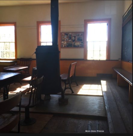 Inside desks and a stove and chalkboards can be seen. The  smooth polished floorboards glow in the afternoon light.